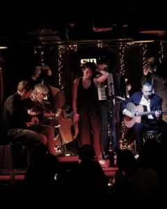 The Tolka Hot Club performing live Gypsy Jazz with a vocalist, violinist, double bassist, and guitarists under warm stage lighting at Annesley House.