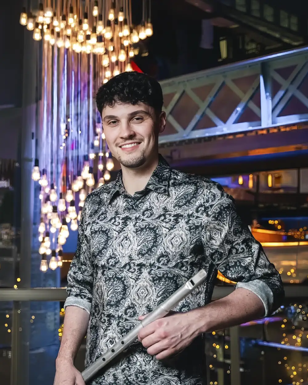 smiling man with short dark hair and a patterned grey and black shirt holds a traditional metal Irish tin whistle, against a background of sparkling hanging bulb lights and a modern balcony.